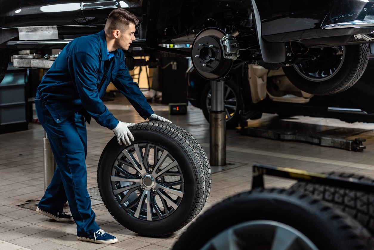 young mechanic holding car wheel near raised car in workshop